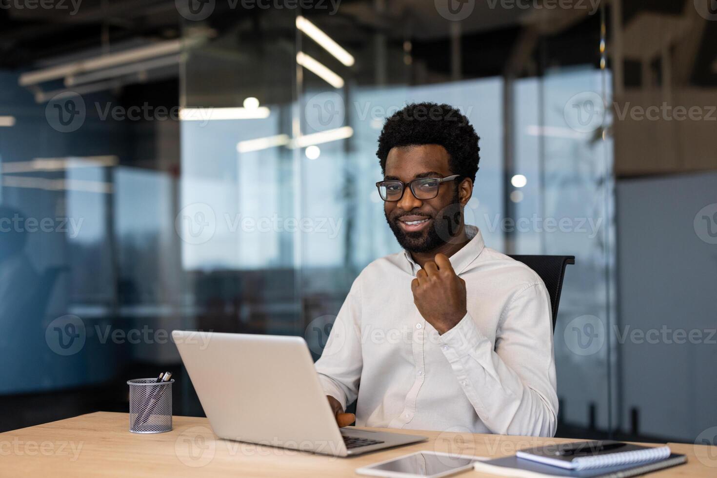 A smiling Black businessman in glasses sits at a desk with a laptop, ready for work. Modern office with glass walls is visible in the background. photo