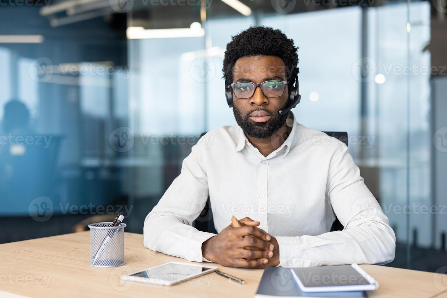 An african-american man wearing a headset sits in an office setting, looking at the camera with a serious expression, ready to assist. photo