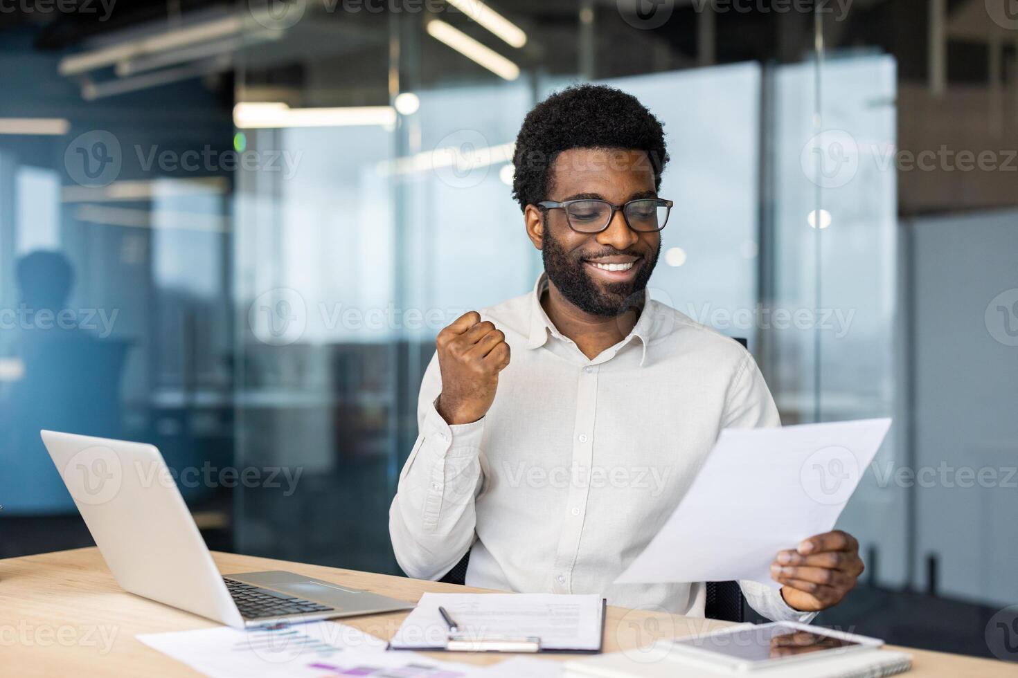 An african american man sits at his desk, ecstatic, holding papers, laptop, and a tablet are on the table. He appears joyful and has an expression of achievement. photo