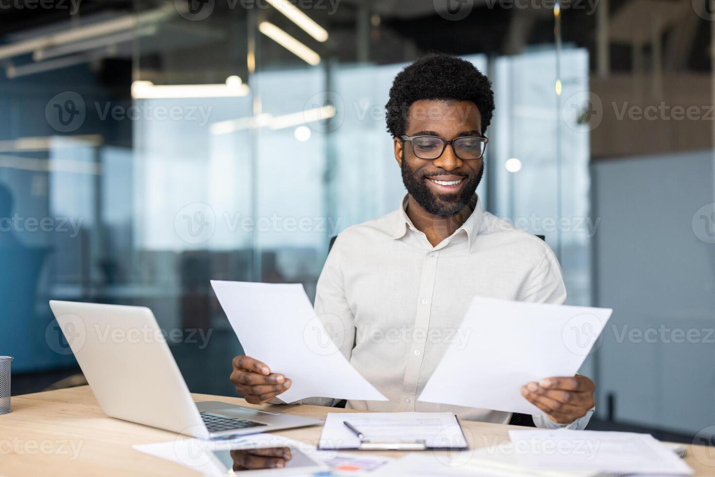 Smiling African American businessman with glasses reviews documents at his desk in a modern office, showcasing a positive work environment photo