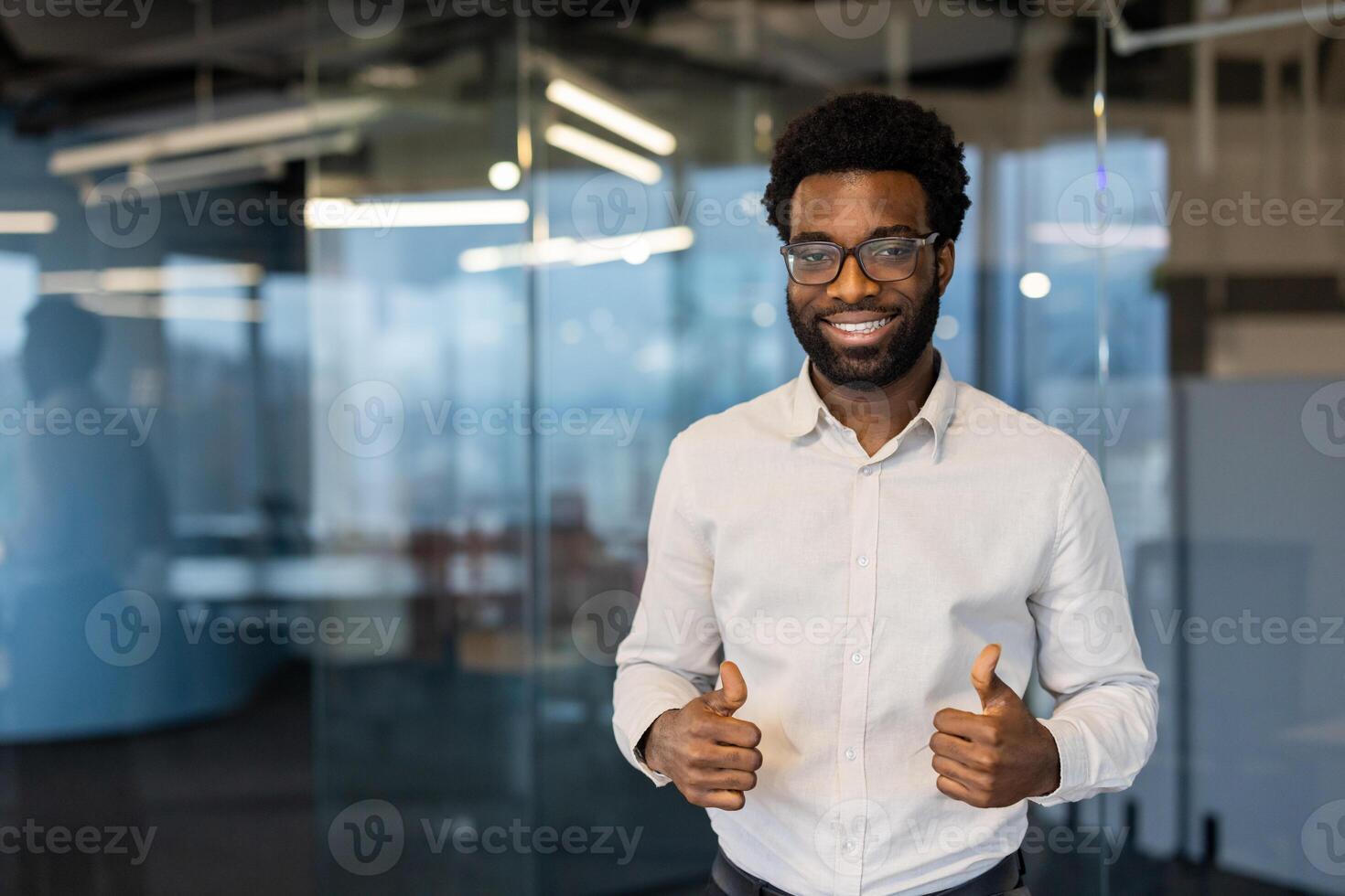 Smiling african american man in eyeglasses gives a thumbs up in an office environment, representing success and approval. photo