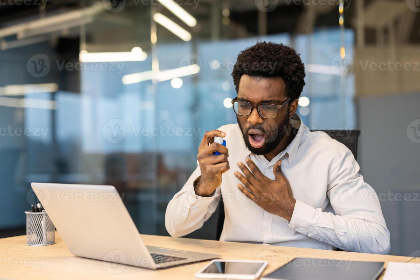An african american man in an office uses an inhaler, potentially experiencing an asthma attack while working on a laptop. photo