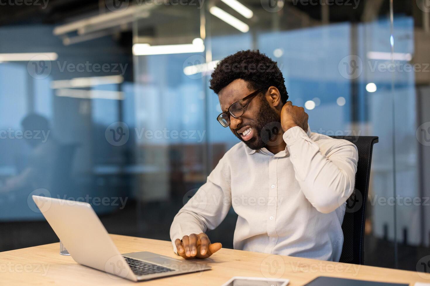 Tired and overworked man suffering from neck pain. Businessman at workplace massaging his neck muscle with his hand, long monotonous sitting work with a laptop inside the office. photo