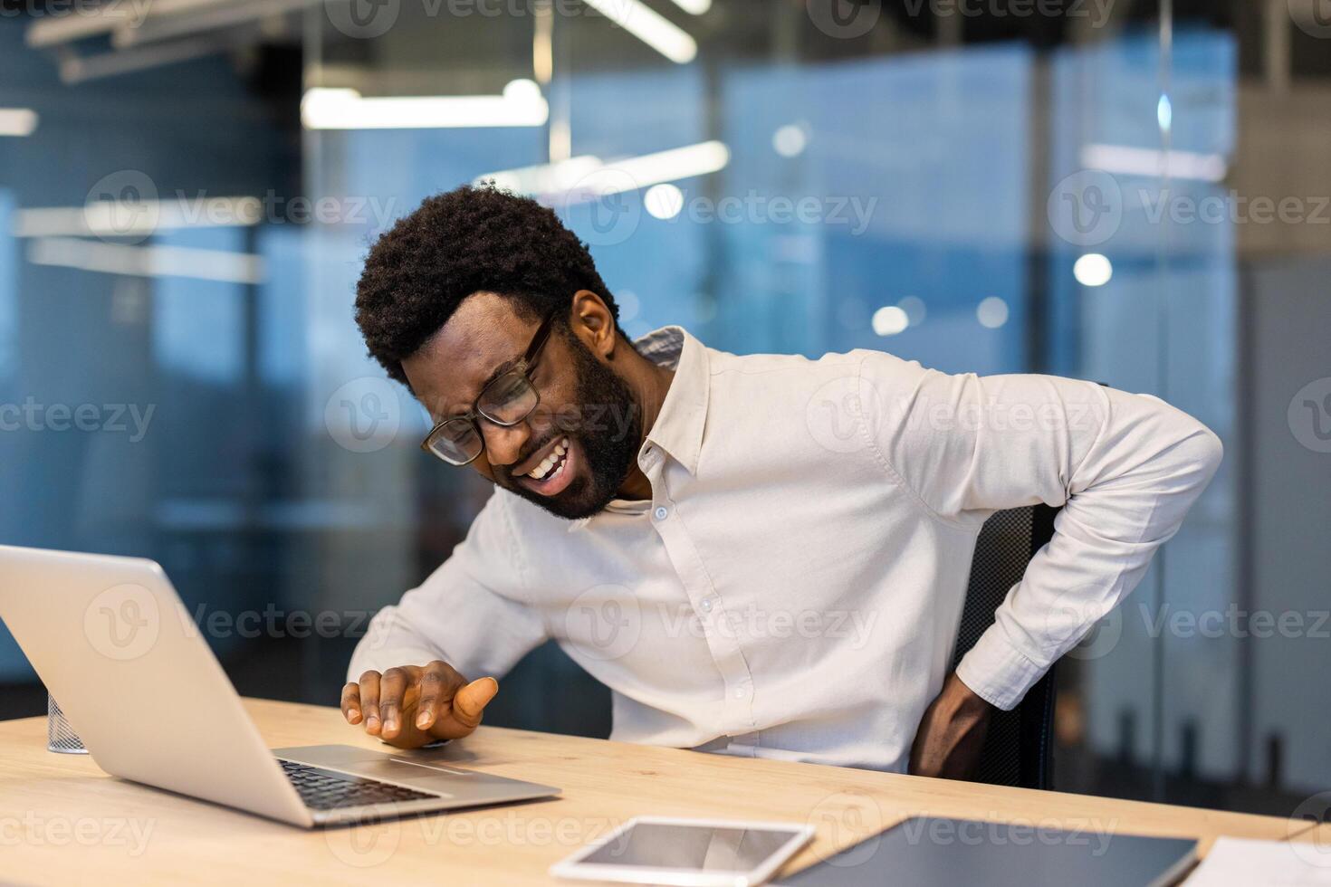 An african american man in glasses experiencing back pain while working on his laptop at the office. He looks stressed. photo