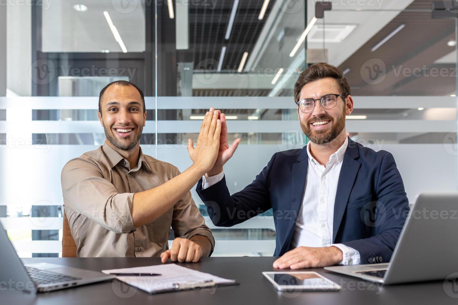 Two diverse business professionals are celebrating success with a high-five gesture in a modern office setting, smiling. photo