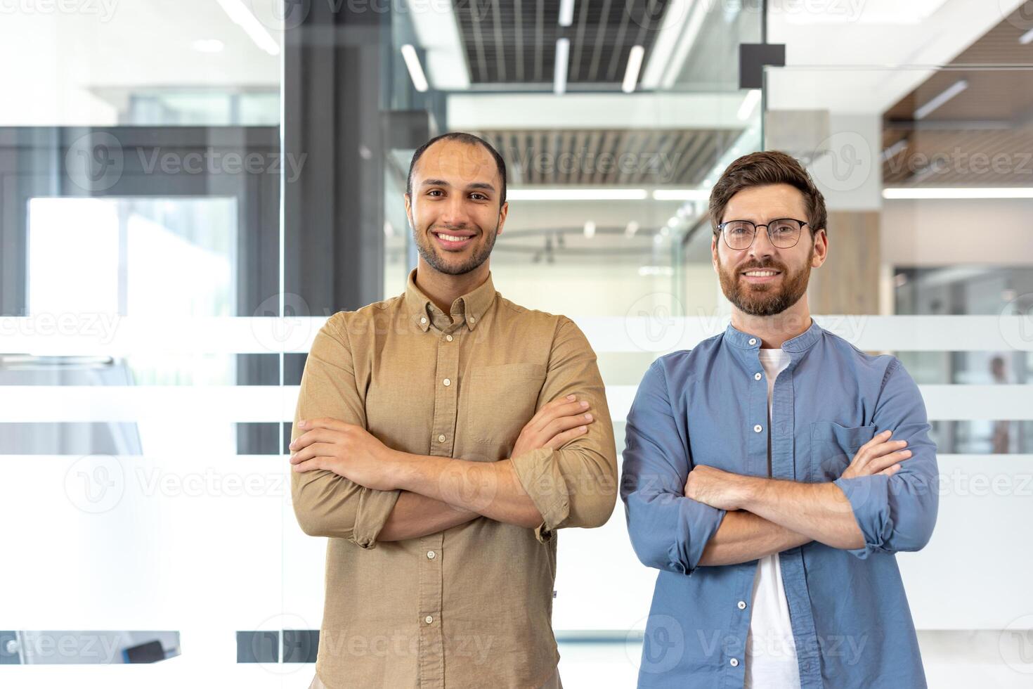 Two confident men stand in an office setting, arms crossed, smiling at the camera. They showcase a professional demeanor. photo