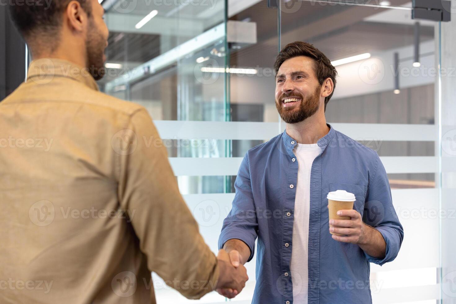 Two business partners shaking hands, smiling, one holding a coffee cup, in a modern office setting. photo