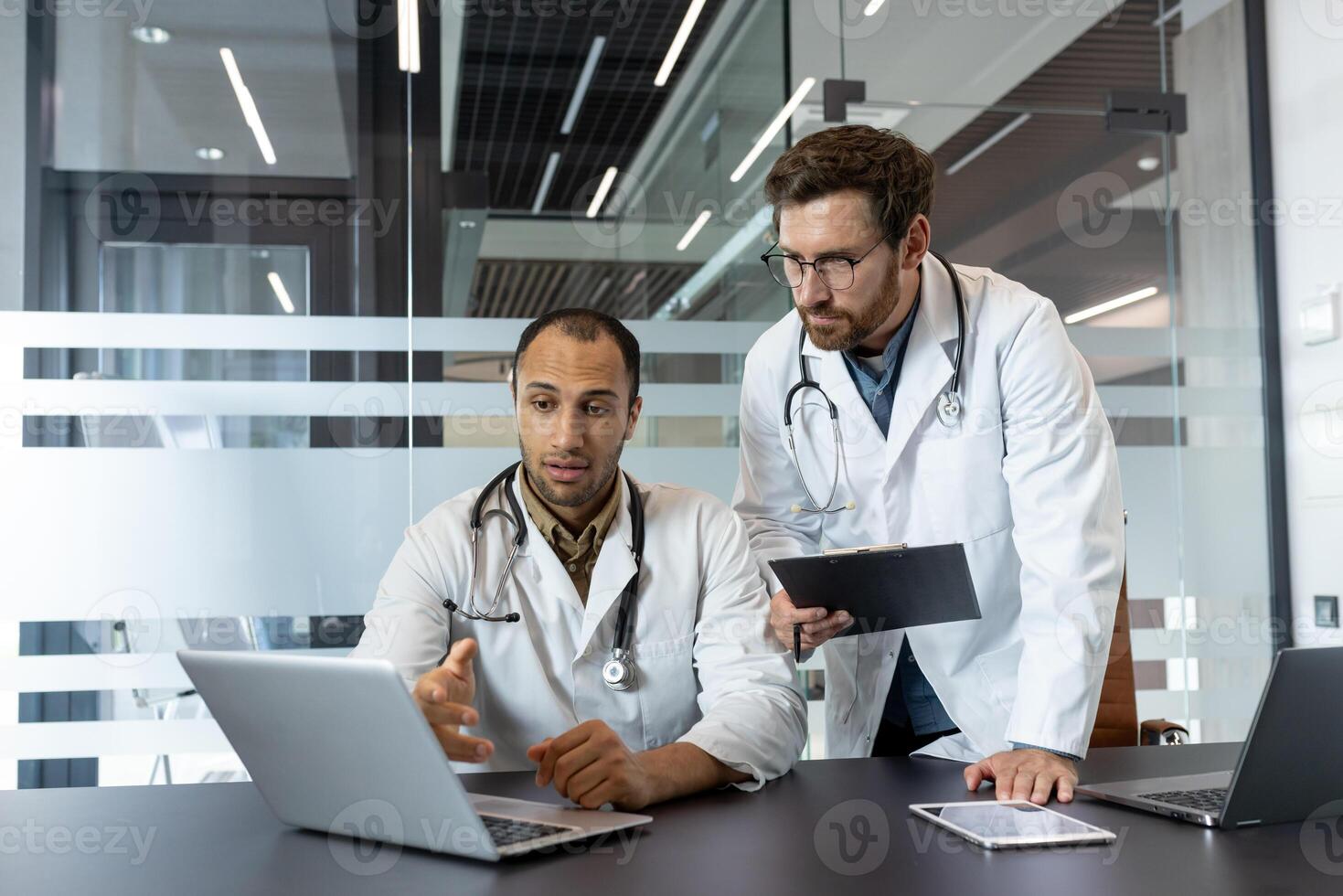 Two doctors in white coats are discussing something on a laptop, a clipboard, and a tablet in a modern office setting. photo