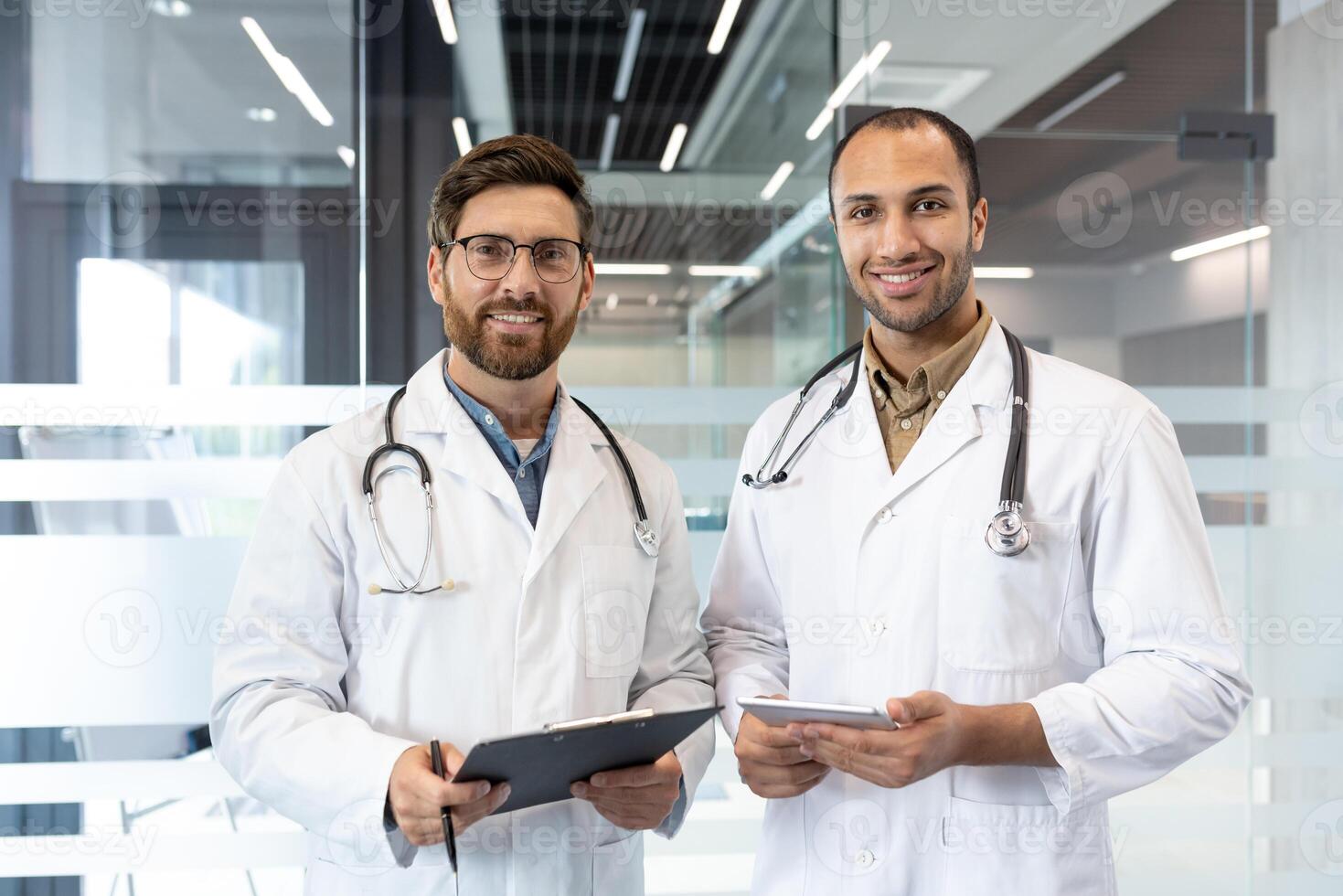 Two doctors in white coats stand side by side, smiling confidently in a modern office setting. One holds a clipboard, the other a tablet. photo