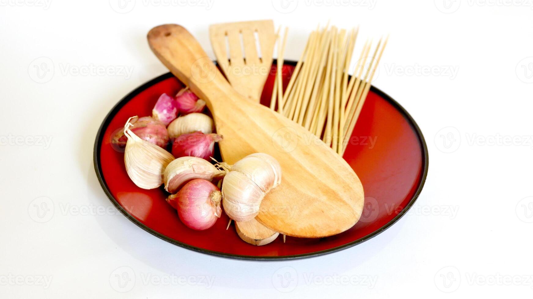 wooden spatula, skewers, and shallots and garlic in a red plastic plate on a white background photo