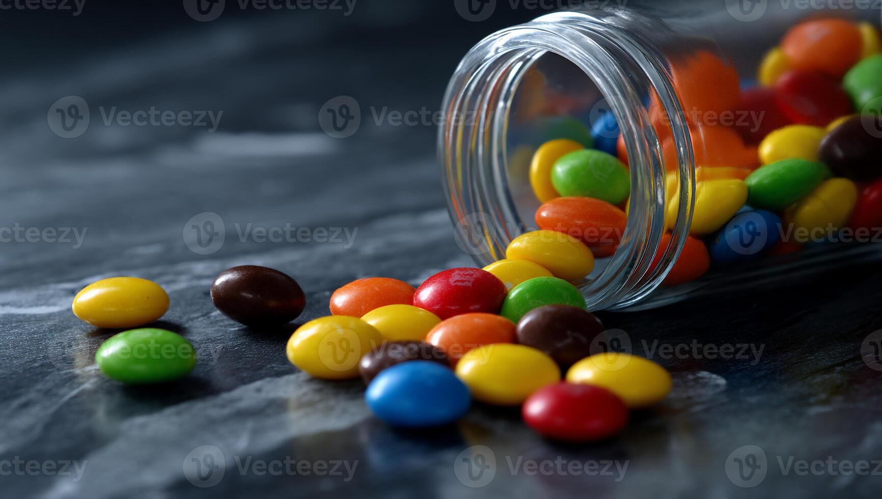 Colorful candies spill from a clear glass jar onto a dark surface photo