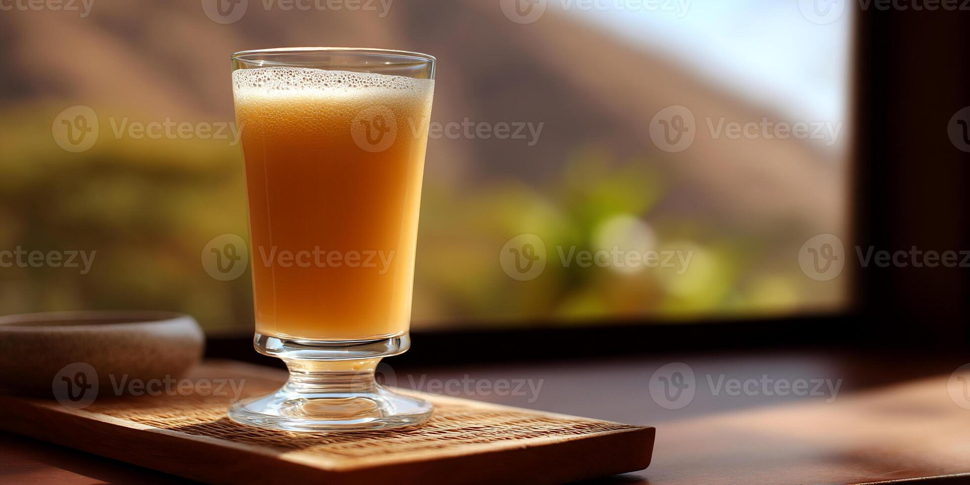 A tall, stemmed glass filled with a light orange smoothie, set on a wooden tray, with a blurred view of a landscape outside a window photo