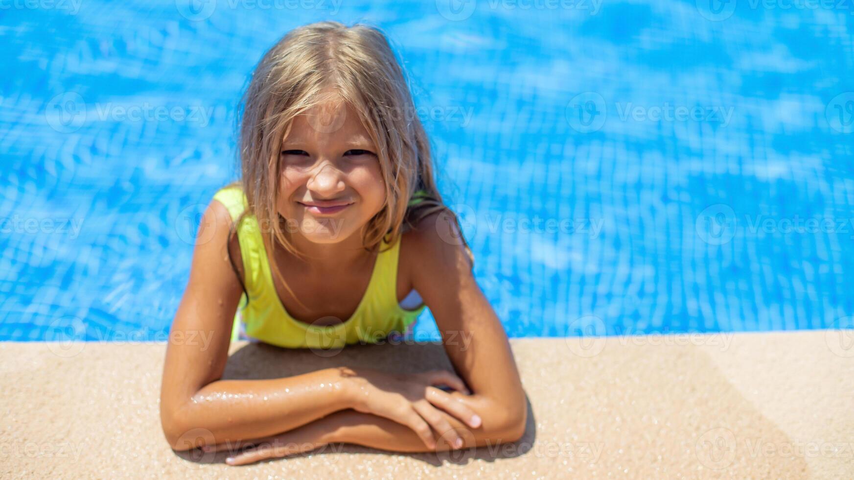 A young girl with blonde hair relaxes at the poolside, leaning on the edge and smiling brightly. The blue water glistens under the sun, capturing a fun summer vibe. photo