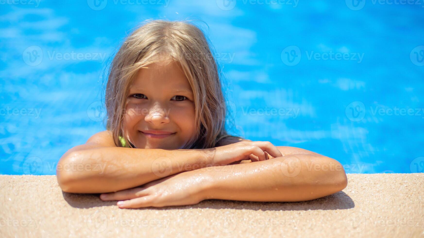 A cheerful child leans on the edge of the pool, smiling with joy as sunlight glimmers on the water. The setting is vibrant and inviting, perfect for a summer day. photo
