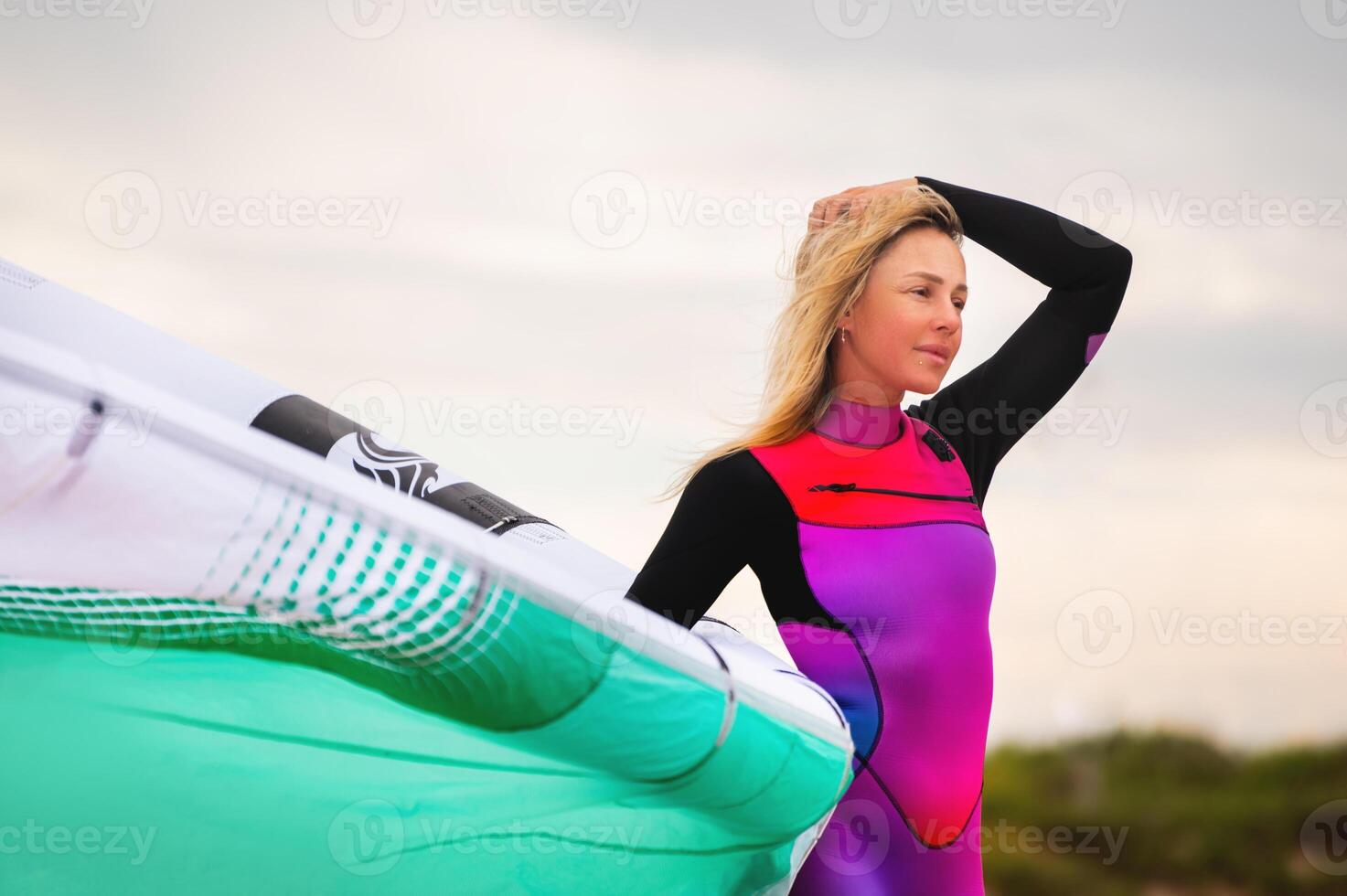 surfer in a wetsuit ponders the weather on a sandy beach while standing in the sand inflating a kite. A sporty girl prepares her equipment before riding photo