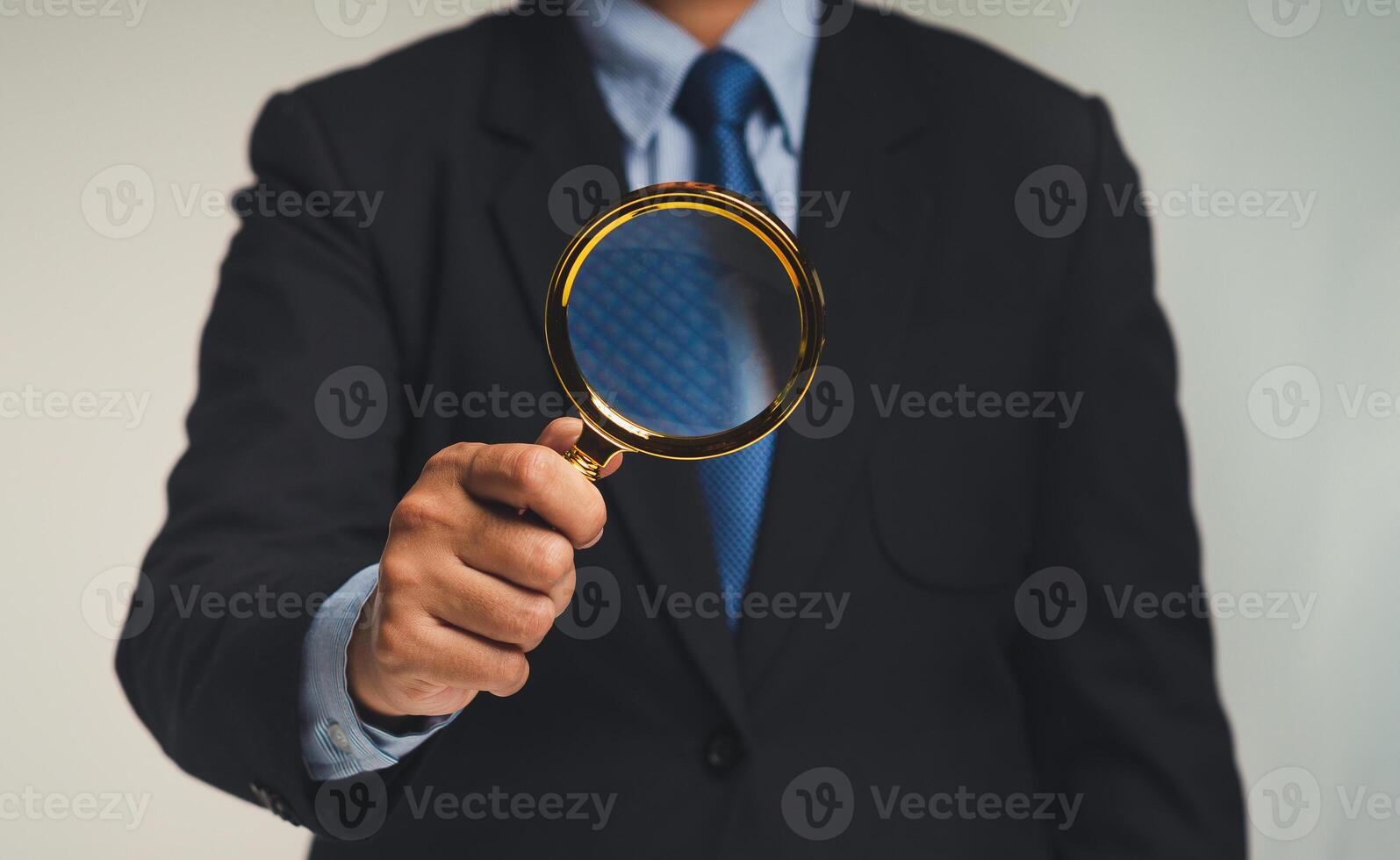 A Businessman in a suit holds a magnifying glass while standing against a black background photo