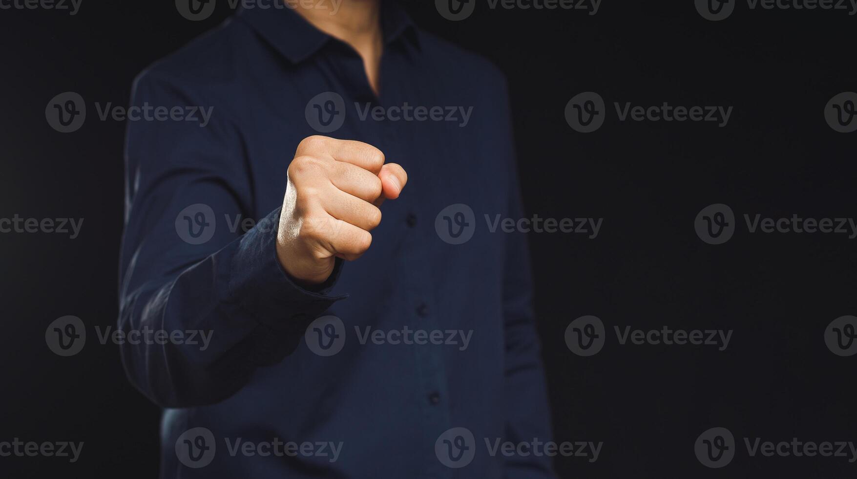 A man in a blue shirt raised his fist while standing against a black background photo