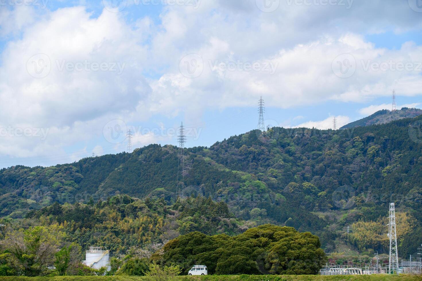 A scenic landscape featuring lush green mountains under a partly cloudy sky. Tall electricity transmission towers line the ridges photo