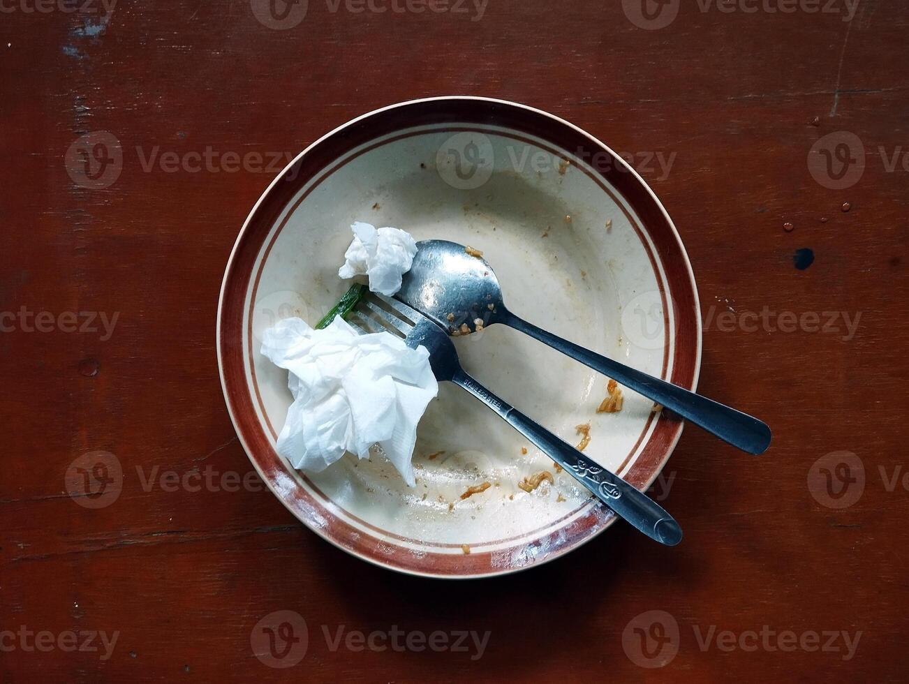 An empty plate with leftover food, a spoon, a fork, and some used tissues placed on a brown table. photo