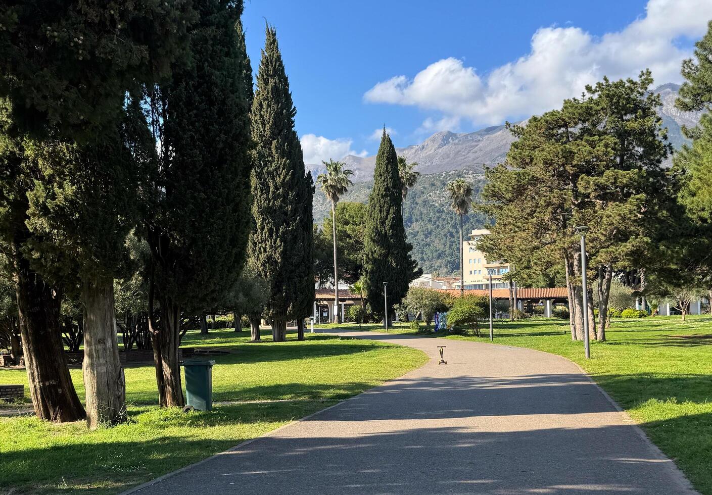 Budva,Montenegro-April 11,2025.A wide paved path winds through a green park with tall cypress and palm trees, and a backdrop of mountains. High quality photo