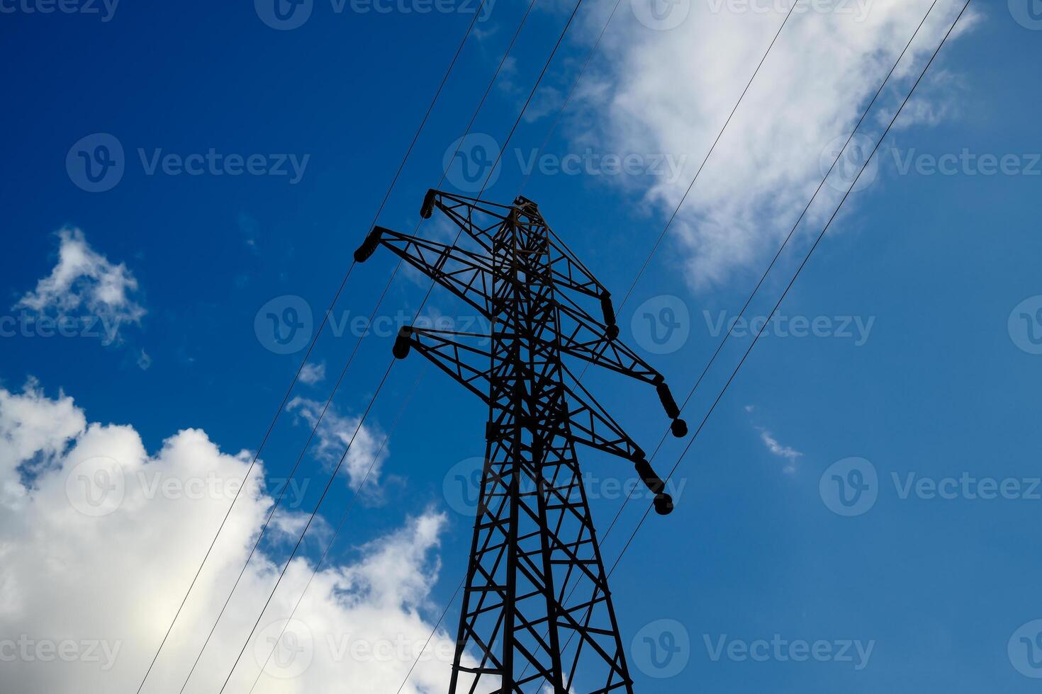 Electrical transmission tower silhouette against bright blue sky with scattered white clouds, high voltage power line pylon photo