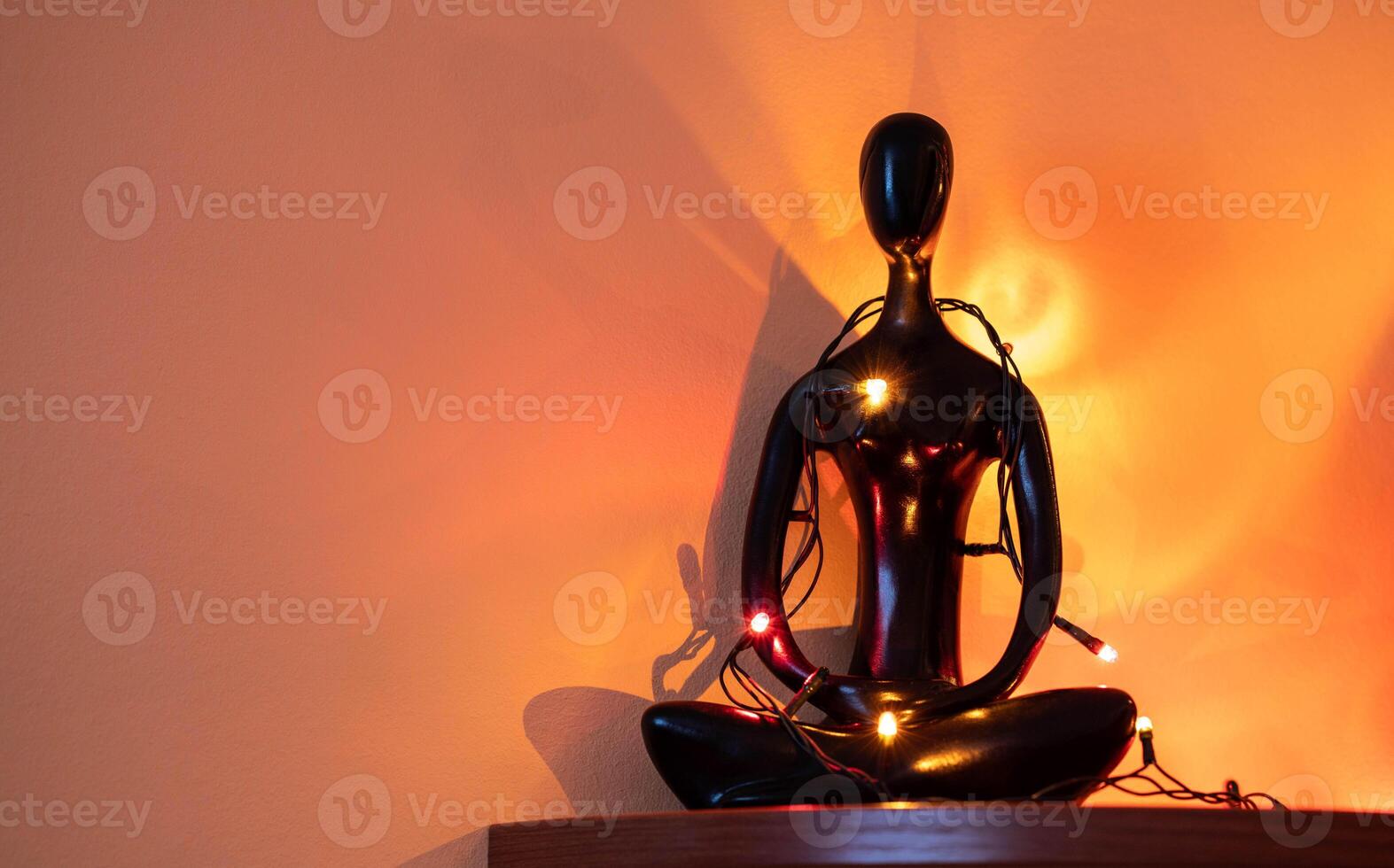 Black figurine of meditating figure adorned with colorful lights against warm wall backdrop photo