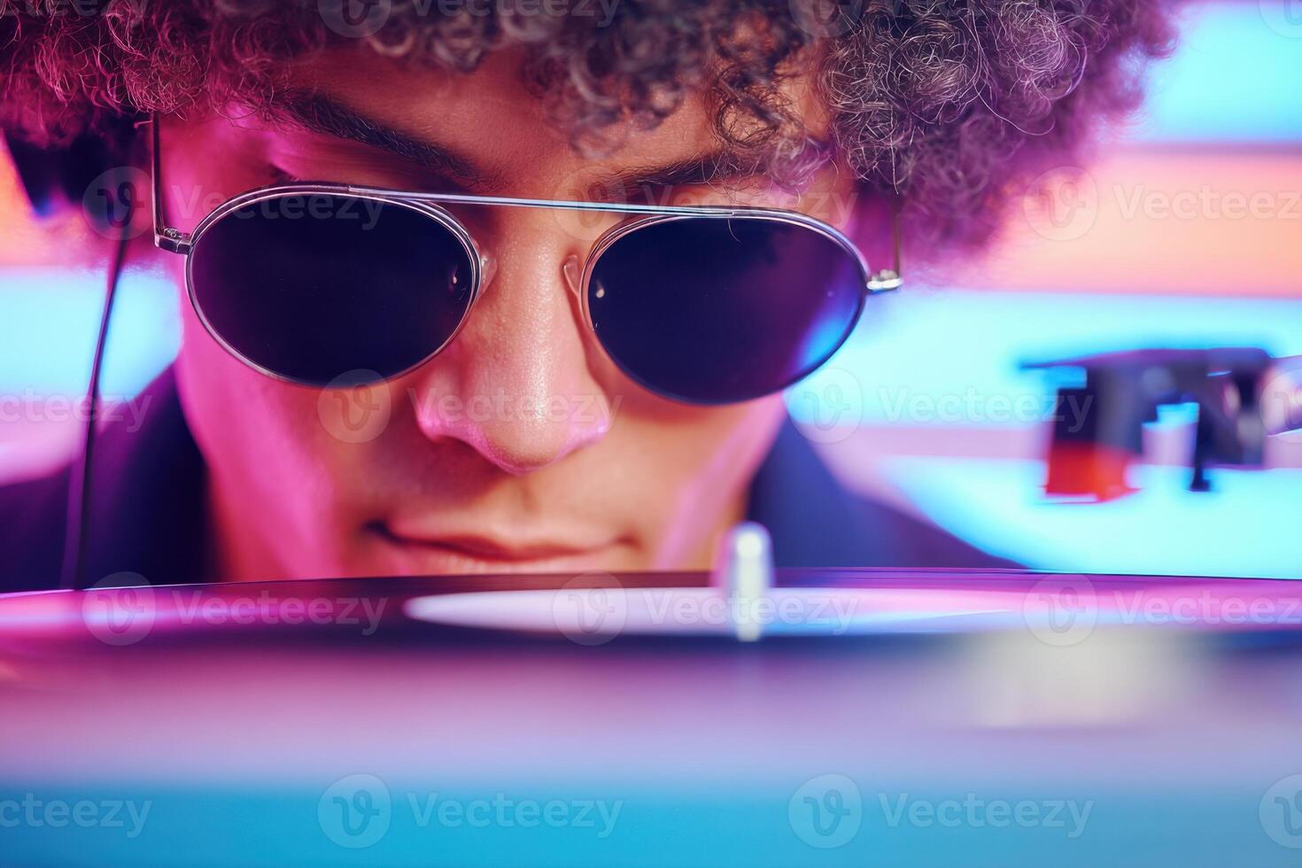 Young man with curly hair wearing sunglasses is focused on a vinyl record player in a vibrant atmosphere, showcasing the art of DJing and music mixing photo