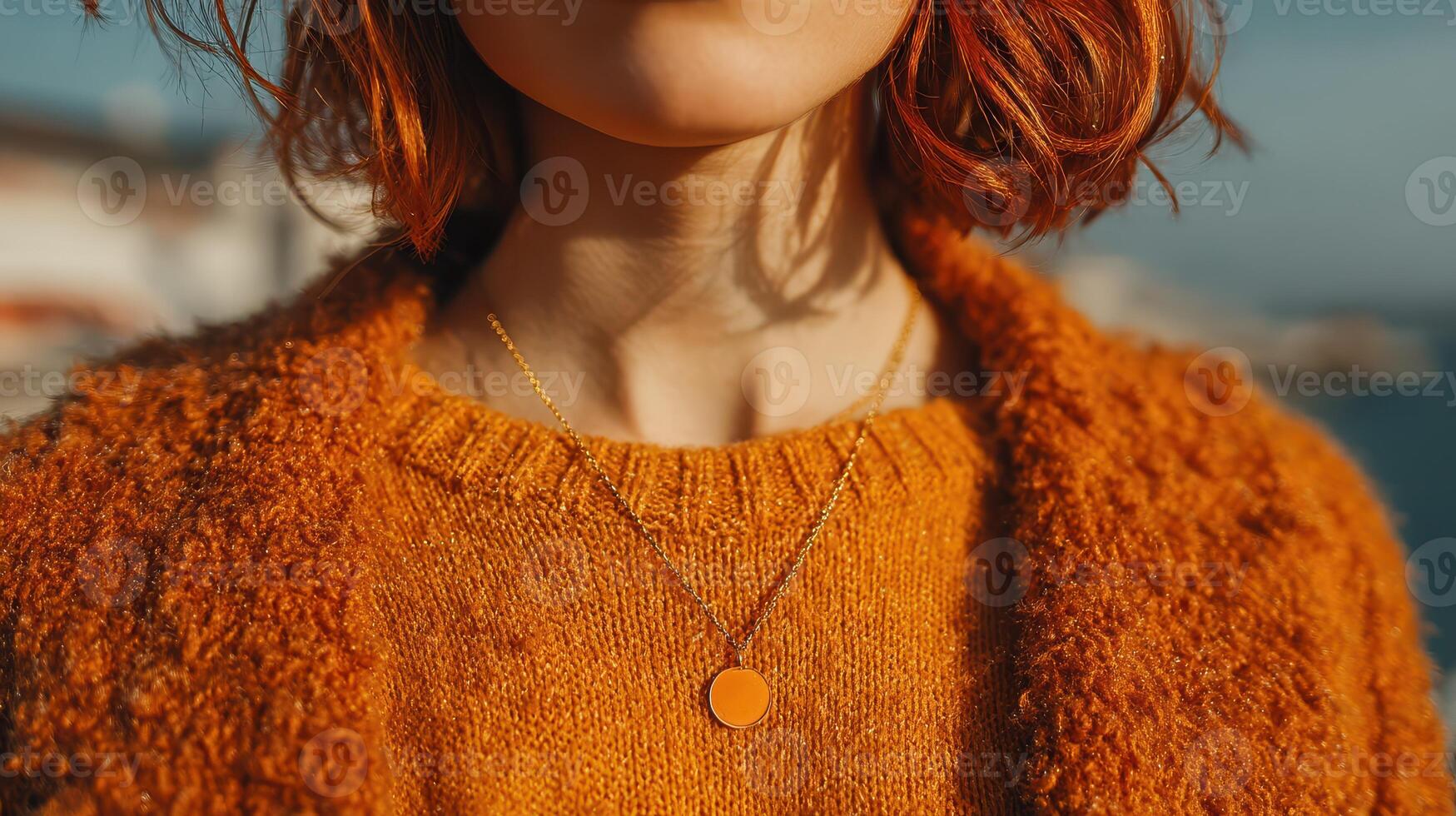 Woman with short red hair wearing an orange sweater and necklace during a sunny day by the water photo