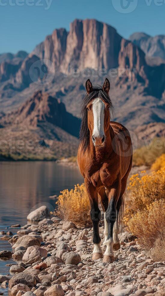 Majestic horse stands along a riverbank with towering mountains in the background under a clear blue sky photo