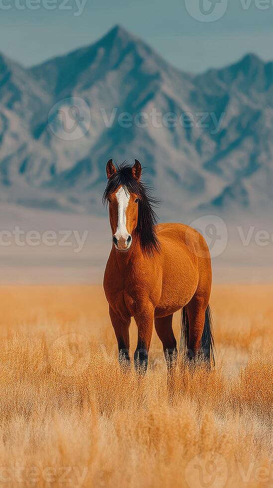 Majestic brown horse standing gracefully in golden grasslands with mountains in the background during a clear day photo