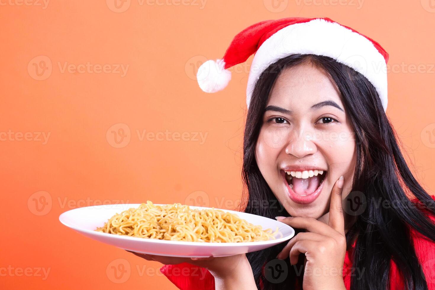 close up portrait of a woman wearing Christmas clothes, Santa hat, happy looking forward, hands holding a white plate filled with noodles to the side and chin, isolated on orange background photo