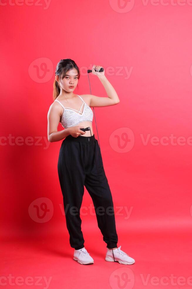 A woman wearing a white top, black pants, and white sneakers, holding a skipping rope, posing for a picture isolated on a red background photo