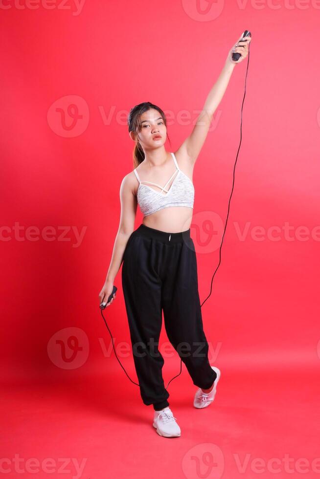 A woman wearing a white top and black pants is holding a jump rope above her head, ready for exercise isolated on a red background photo