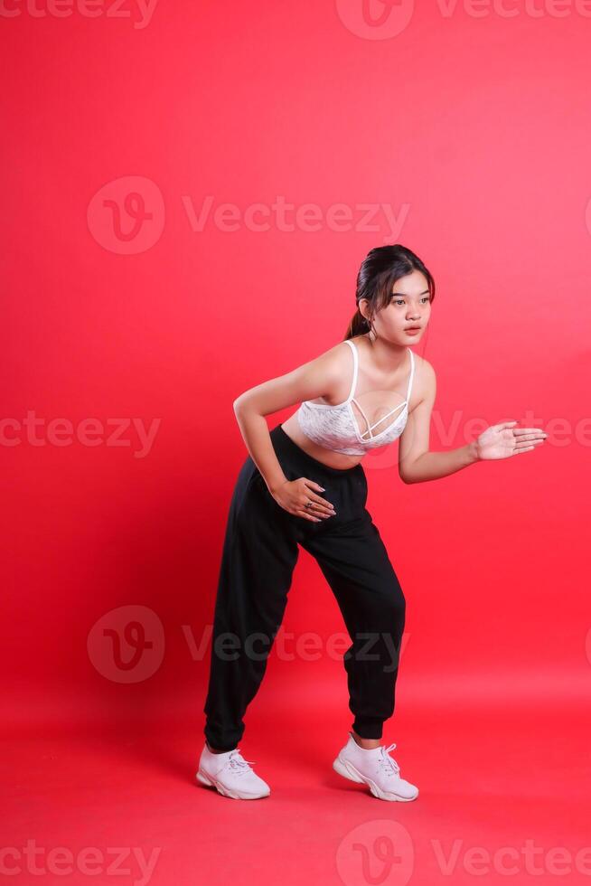 A woman in a white sports bra and black pants, with white sneakers is seen posing with arms extended. isolated on a red background photo