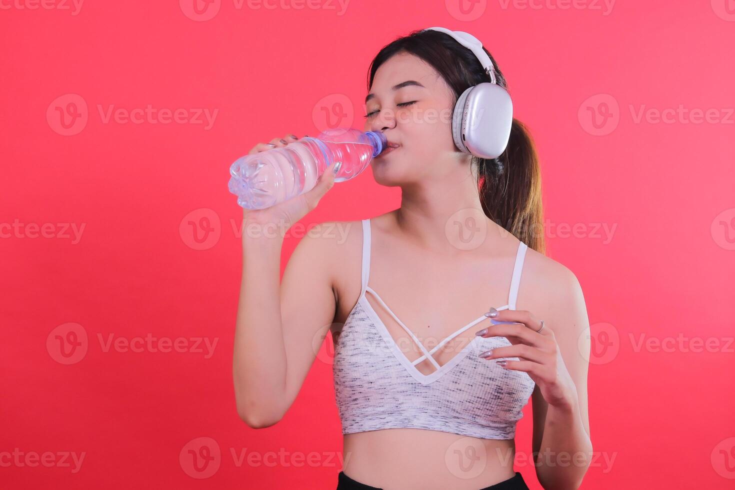 A woman in athletic attire drinks from a clear water bottle, wearing headphones, with her eyes closed, isolated on a red background. photo