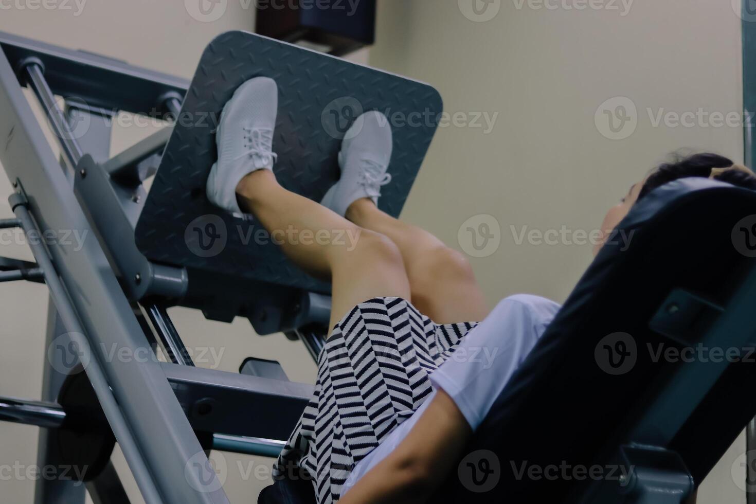 women using the leg press machine are wearing white shoes, shorts and a white shirt. Their feet are positioned on the gray textured foot bed of the machine. The leg press machine is made of gray metal photo