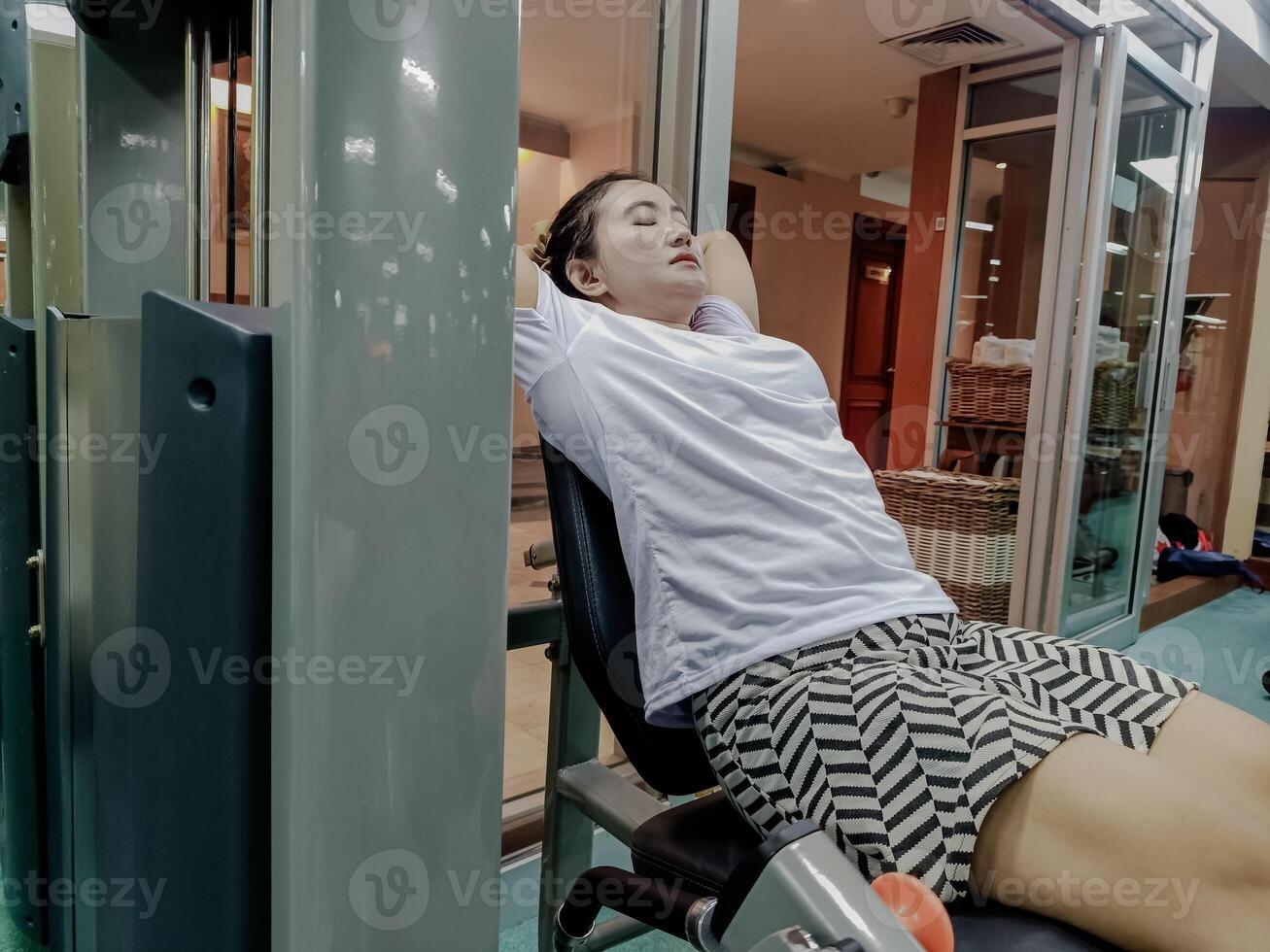 a woman in a white shirt and patterned shorts. Fitness equipment is visible from the inside view of the fitness center, closing her eyes and putting her arms behind her head photo