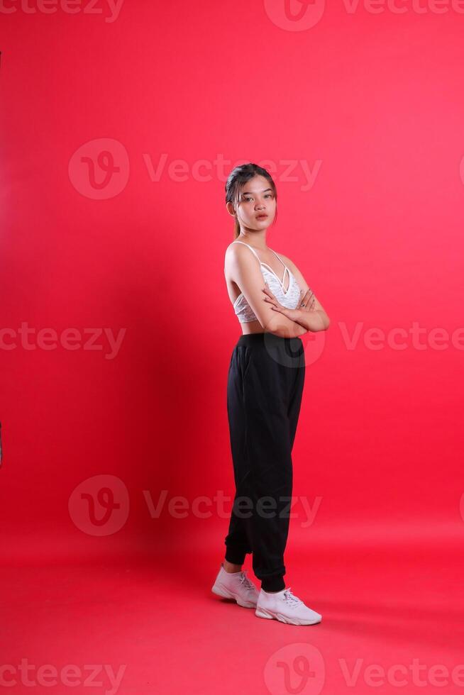 A young woman dressed in white top and black pants stands with her arms crossed looking at the camera, isolated on a red background photo