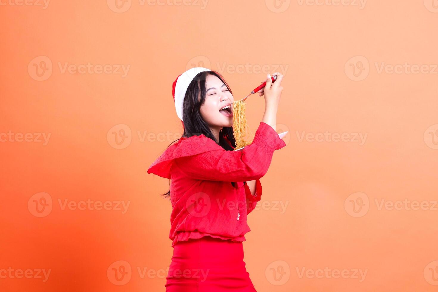 gesture of beautiful woman wearing red christmas blouse, Santa hat, left slant enjoying eating noodles, hand holding noodle fork and white plate filled with noodles, isolated on orange background photo