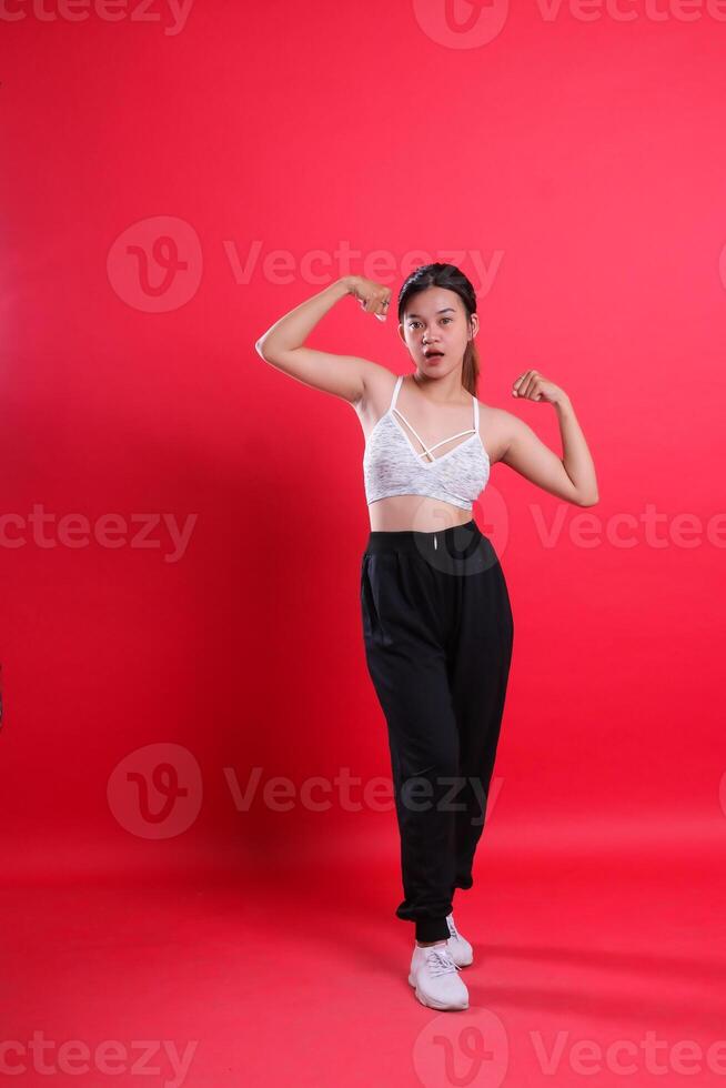A woman in a white top, black pants and white shoes flexes her arm muscles, isolated on a red background. photo