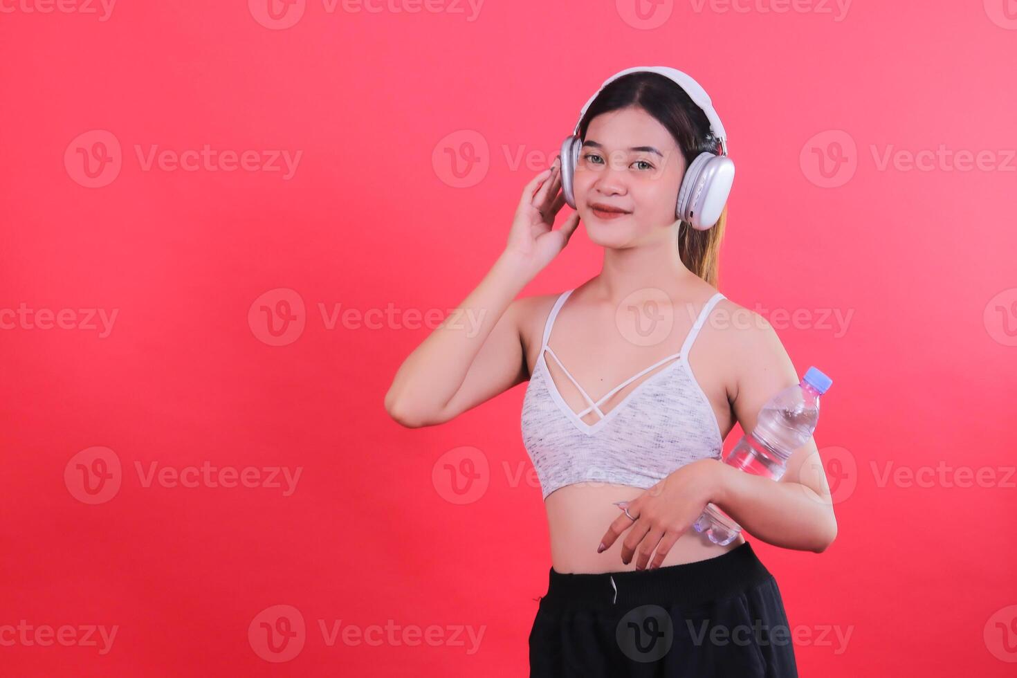 A woman wearing sport bra, black shorts and headphones holds a water bottle in her hands, isolated on a red background photo