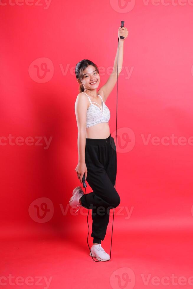 A woman in fitness attire is holding a jump rope, standing on one leg, isolated on a red background. photo