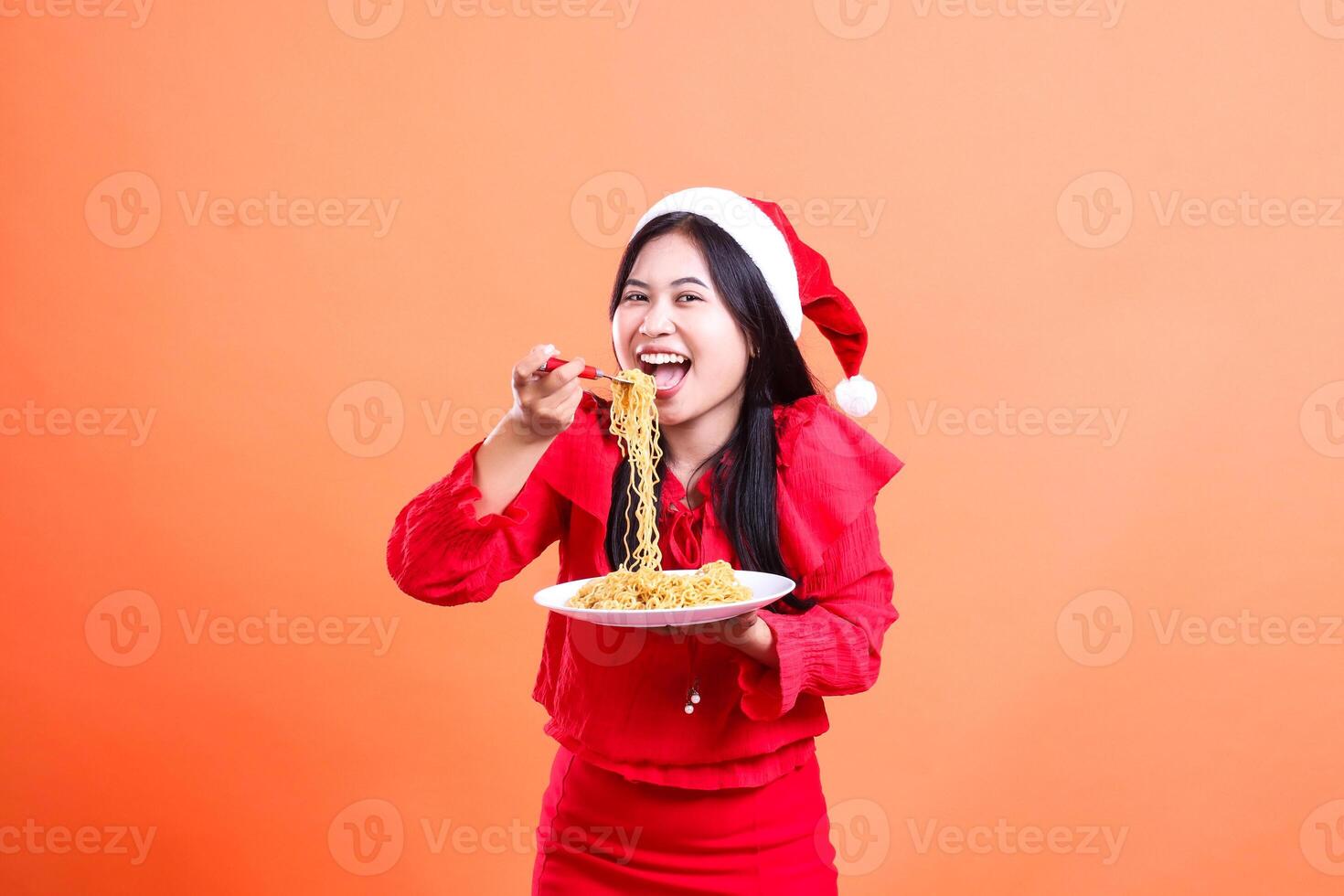 Asian young woman beauty cheerful looking at camera, hand holding fork filled with noodles from white plate while eating it, wearing red christmas blouse, isolated on orange background photo