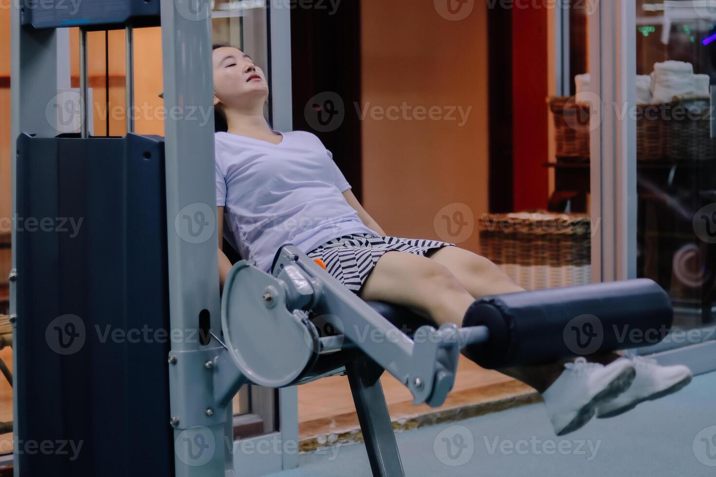 A woman asia uses a leg extension machine at a fitness center. The woman sits with her legs stretched out on the padded bar of the machine. She is wearing a white shirt and shorts. photo