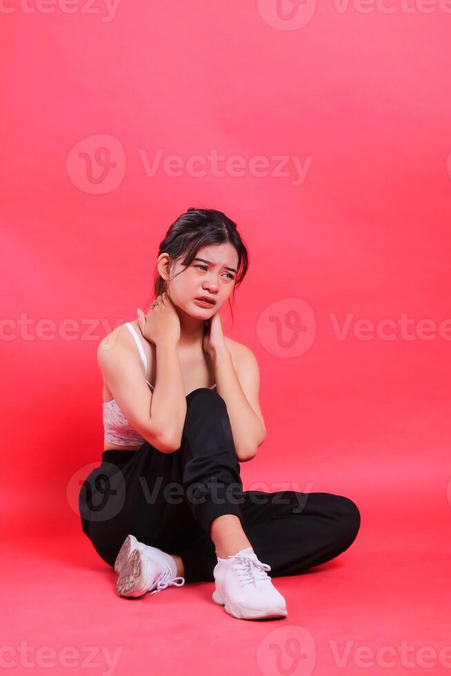 A woman asia sits holding her sore and painful neck while wearing black sneakers and pants, isolated on a red background. photo