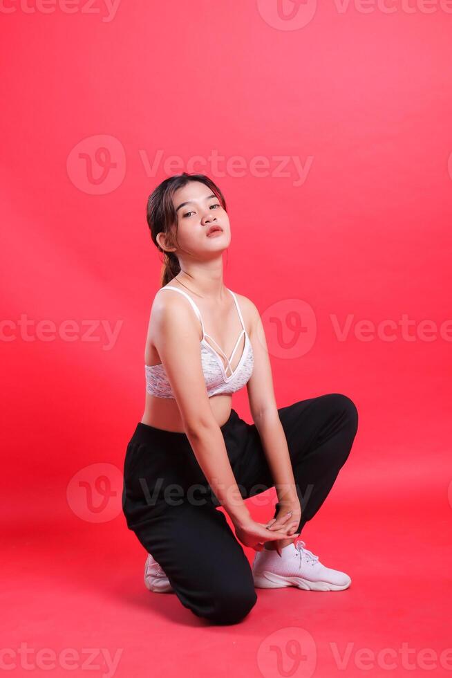 A young woman wearing sports bra, pants and white sneakers, poses while crouching, isolated on a red background. photo