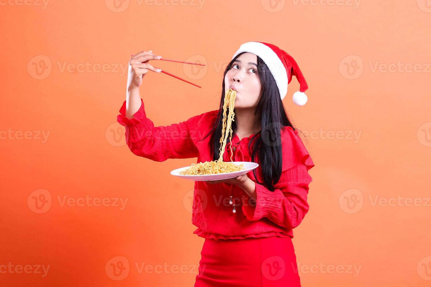 young indonesia woman shocked f slurping noodles , hands holding chopsticks and white plate filled with noodles , wearing red christmas blouse , Santa hat , isolated on orange background photo