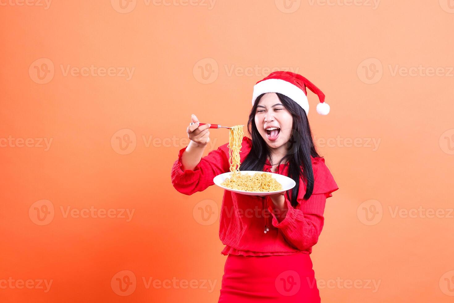portrait of a girl wearing a Christmas dress, Santa hat, screaming looking at noodles, hand holding a noodle fork and a white plate filled with noodles in front, isolated on an orange background photo