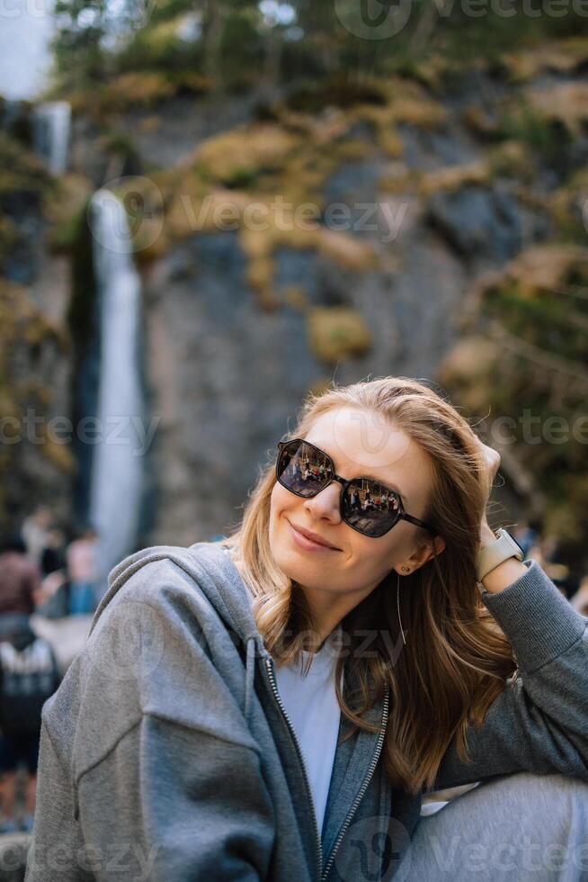 Young blonde woman wearing sunglasses and casual clothing sitting and smiling in front of waterfall in mountain valley while enjoying landscape. Blonde woman in casual attire radiating happiness photo