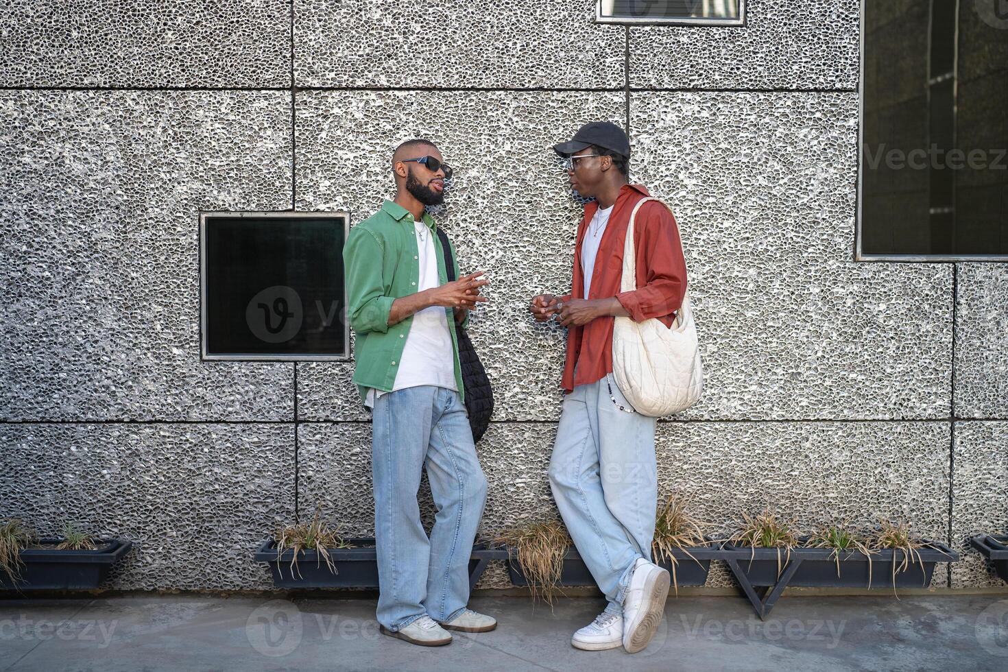 African American student duo engages in lively chat near wall on city walk. Stylish modern youth photo